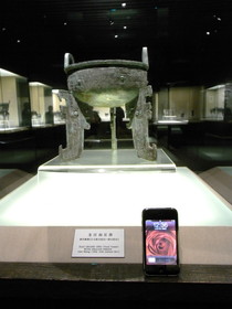 Tomato Juice‘s China Travel とまとじゅーす的中国旅行、上海博物館・青銅器展。先ほどの商代晩期、紀元前13～11世紀頃の龍紋扁足鼎という青銅器とiPhoneの大きさを比較